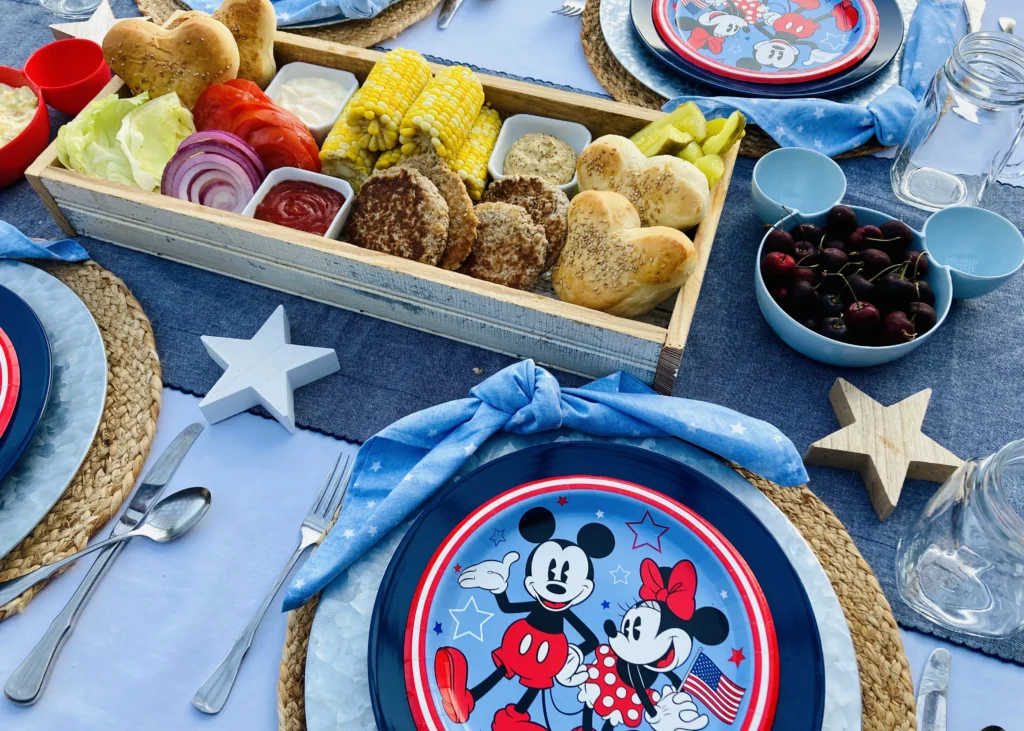 Mickey Burger Board with Mickey-shaped buns, burger patties, corn on the cob, lettuce, tomato, red onion, pickles, mayo, and mustard arranged on a long farmhouse tray for a Disney Patriotic Farmhouse BBQ