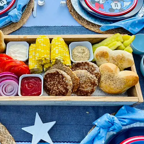 Mickey Burger Board with Mickey-shaped buns, burger patties, corn on the cob, lettuce, tomato, red onion, pickles, mayo, and mustard arranged on a long farmhouse tray for a Disney Patriotic Farmhouse BBQ