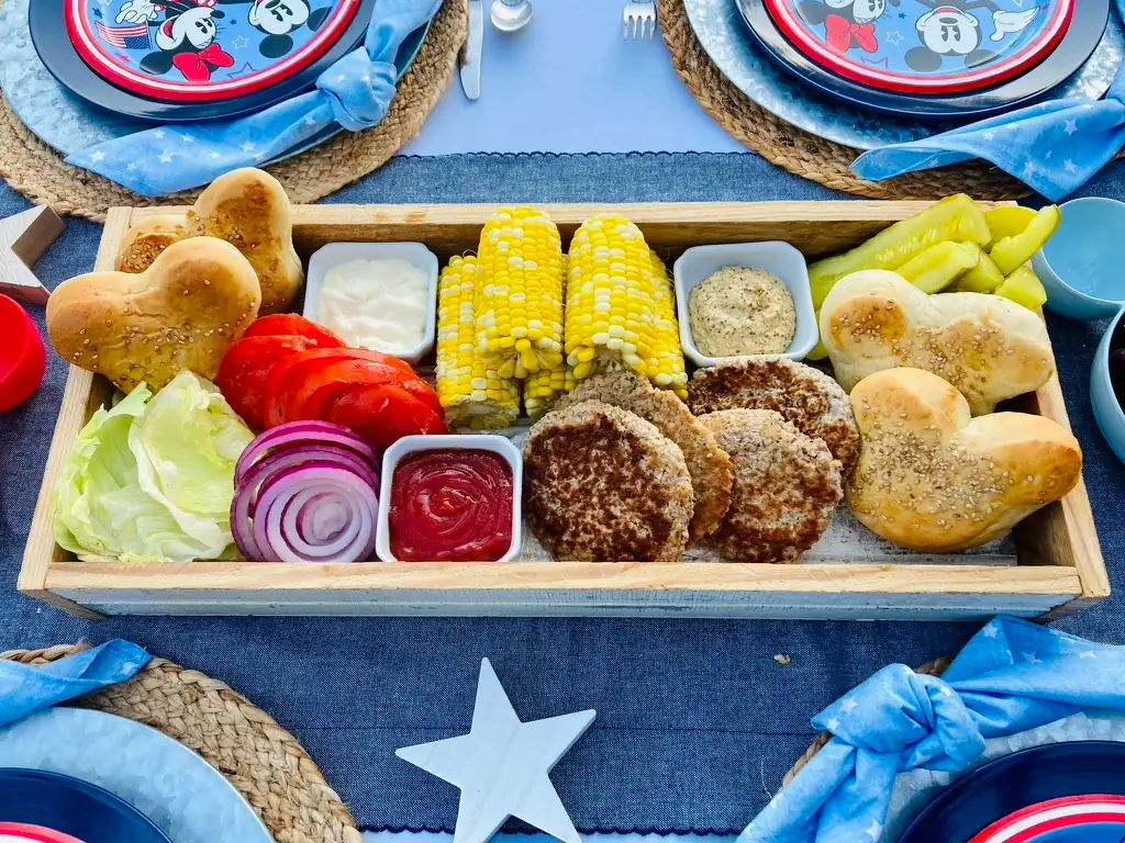 Mickey Burger Board with Mickey-shaped buns, burger patties, corn on the cob, lettuce, tomato, red onion, pickles, mayo, and mustard arranged on a long farmhouse tray for a Disney Patriotic Farmhouse BBQ