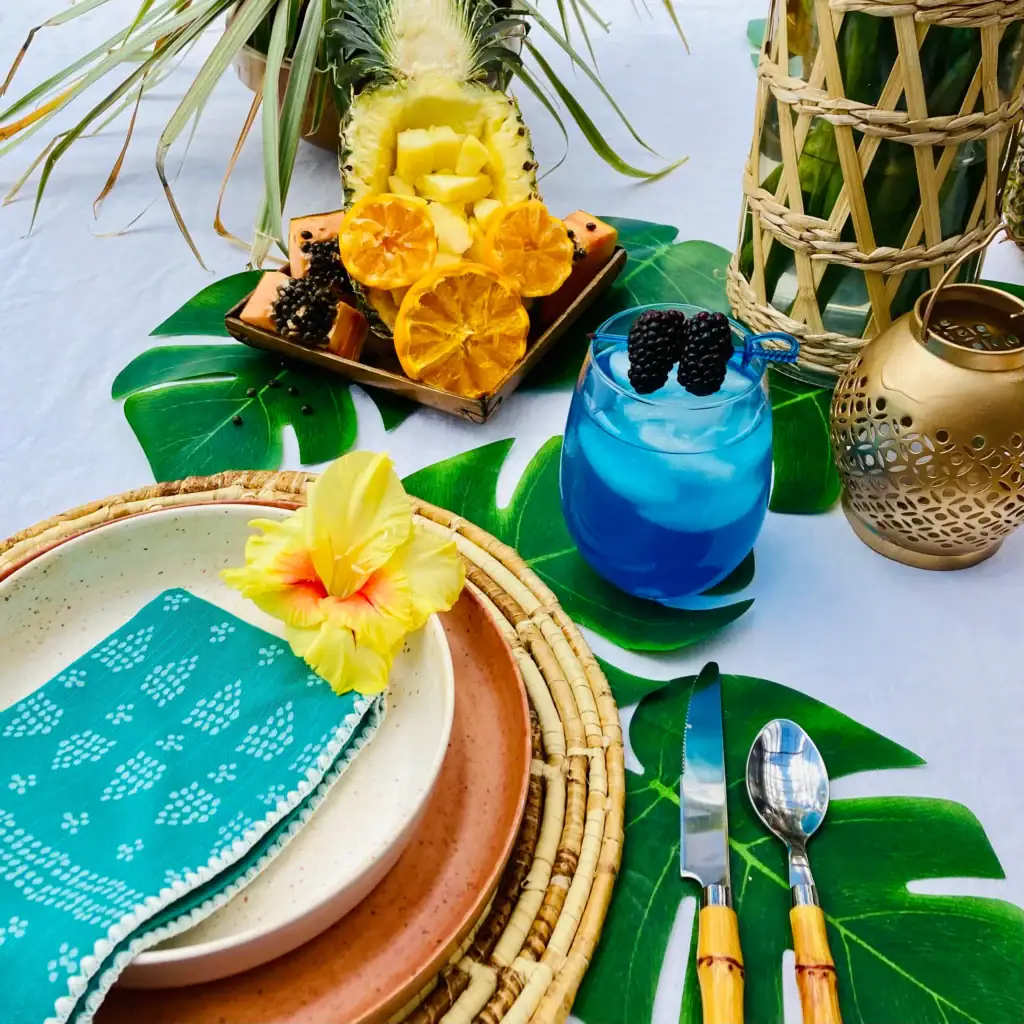 Aladdin's Blue Calypso Cooler mocktail in stemless glasses with vivid blue Calypso lemonade, ice, and fresh blackberry garnish on blue cocktail picks, styled on a tropical table with monstera leaves and woven placemats for an Enchanted Tiki Room Disney brunch.