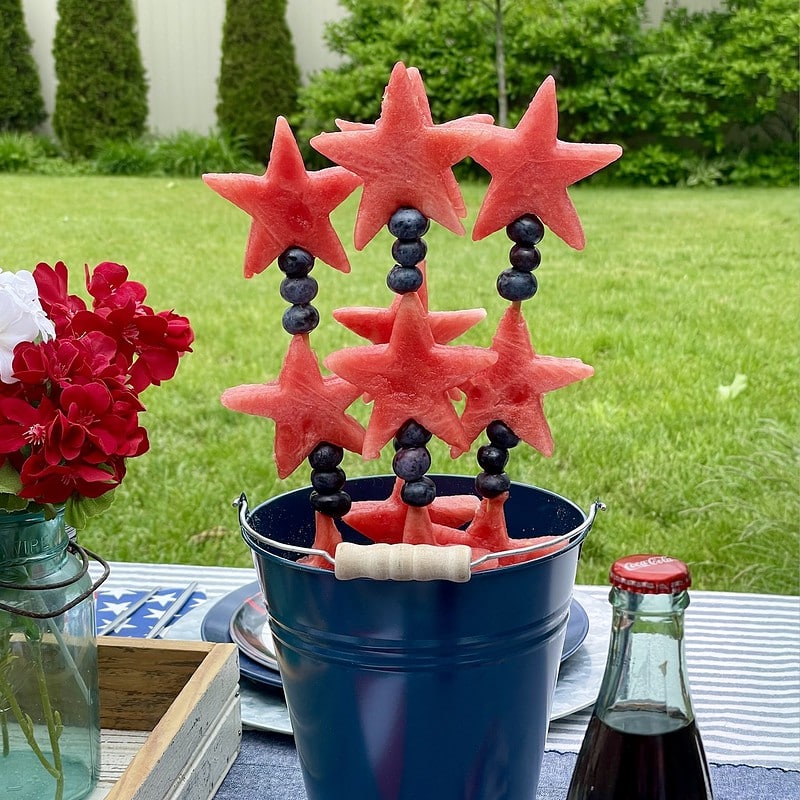 Stars & Stripes watermelon fruit kabobs with watermelon stars and fresh blueberries on bamboo skewers, displayed upright in a navy metal bucket on a patriotic Captain America party table outdoors.