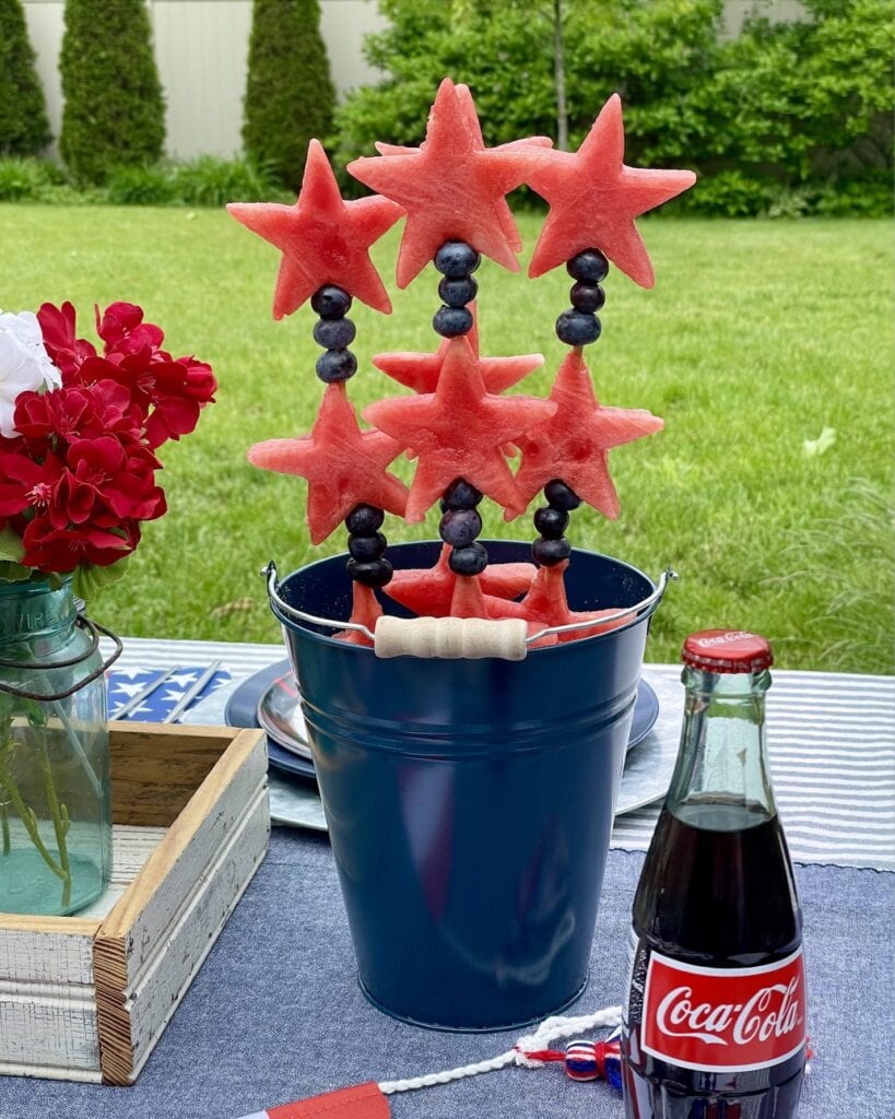 Stars & Stripes watermelon fruit kabobs with watermelon stars and fresh blueberries on bamboo skewers, displayed upright in a navy metal bucket on a patriotic Captain America party table outdoors.