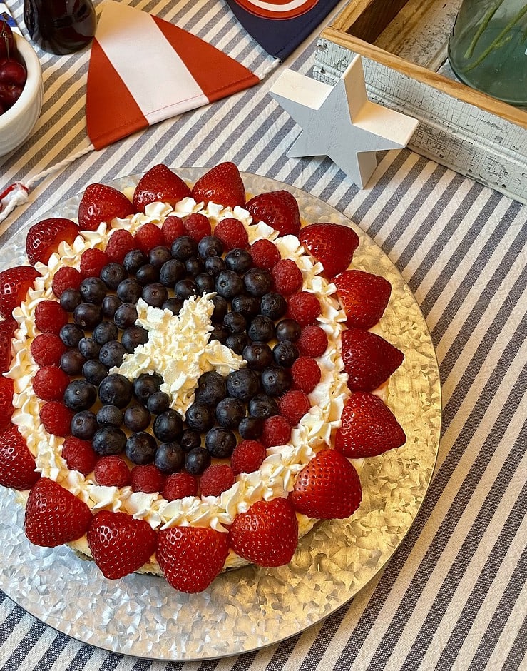 Captain America Shield Cheesecake decorated with concentric rings of fresh strawberries, raspberries, blueberries, and piped whipped cream stars to recreate the Captain America shield, styled on a striped tablecloth with Captain America party decor.