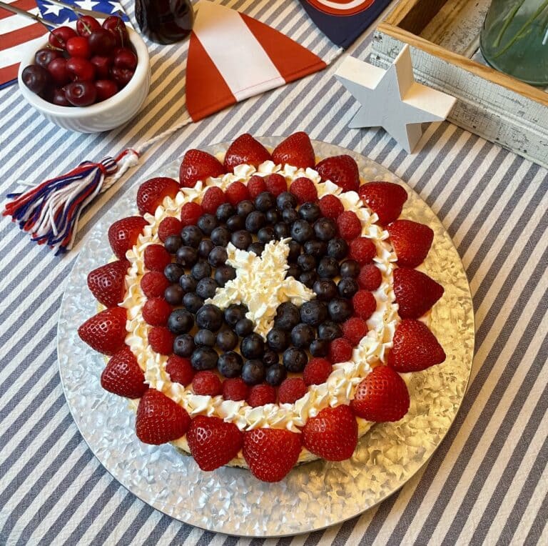 Captain America Shield Cheesecake decorated with concentric rings of fresh strawberries, raspberries, blueberries, and piped whipped cream stars to recreate the Captain America shield, styled on a striped tablecloth with Captain America party decor.
