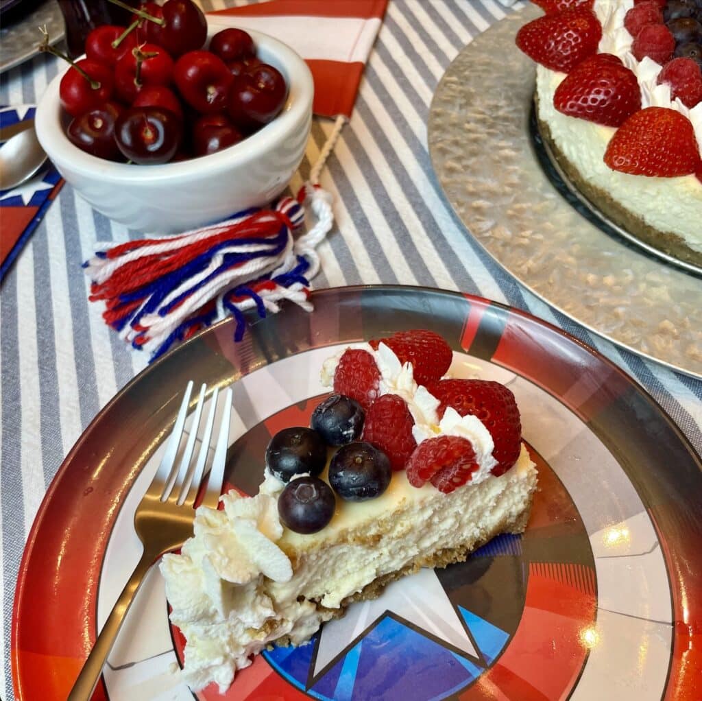 Captain America Shield Cheesecake decorated with concentric rings of fresh strawberries, raspberries, blueberries, and piped whipped cream stars to recreate the Captain America shield, styled on a striped tablecloth with Captain America party decor.