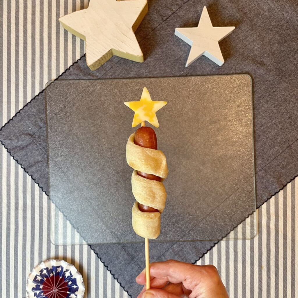Golden breadstick-wrapped hot dog on a bamboo skewer with a yellow cheese star on top, styled for a 4th of July Captain America cookout.