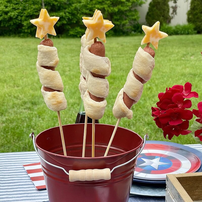 Captain America Rocket Dogs — breadstick-wrapped hot dogs on bamboo skewers topped with yellow cheese stars, displayed upright in a red metal bucket on a patriotic Captain America party table with a blue striped tablecloth.