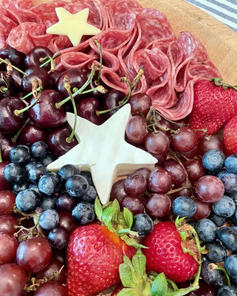 Captain America star charcuterie board on a round wood tray with star-shaped brie filled with raspberry jam, folded salami, white cheese stars, fresh strawberries, blueberries, cherries, and red grapes for a 4th of July Marvel party.