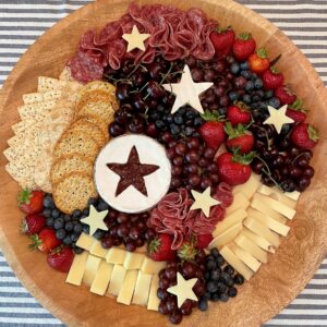 Captain America star charcuterie board on a round wood tray with star-shaped brie filled with raspberry jam, folded salami, white cheese stars, fresh strawberries, blueberries, cherries, and red grapes for a 4th of July Marvel party.