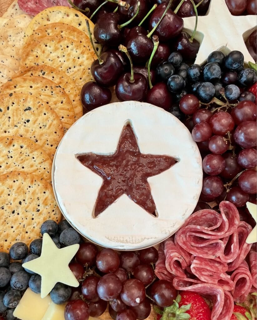 Captain America star charcuterie board on a round wood tray with star-shaped brie filled with raspberry jam, folded salami, white cheese stars, fresh strawberries, blueberries, cherries, and red grapes for a 4th of July Marvel party.