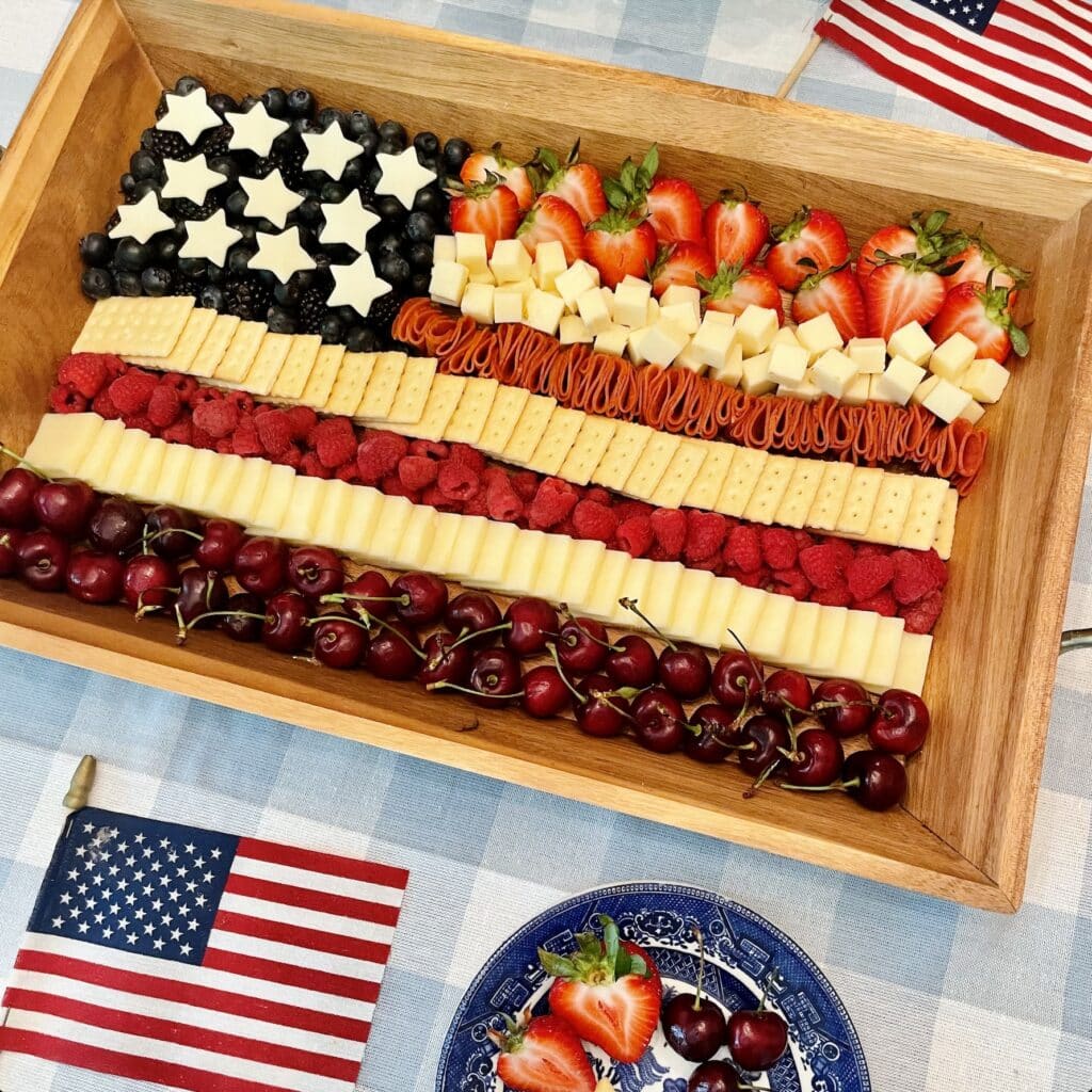 Flag Charcuterie Board on a wooden tray with red, white, and blue stripes of cherries, cheese, crackers, and berries arranged to look like the American flag, styled with white hydrangeas and American flags for a 4th of July celebration.