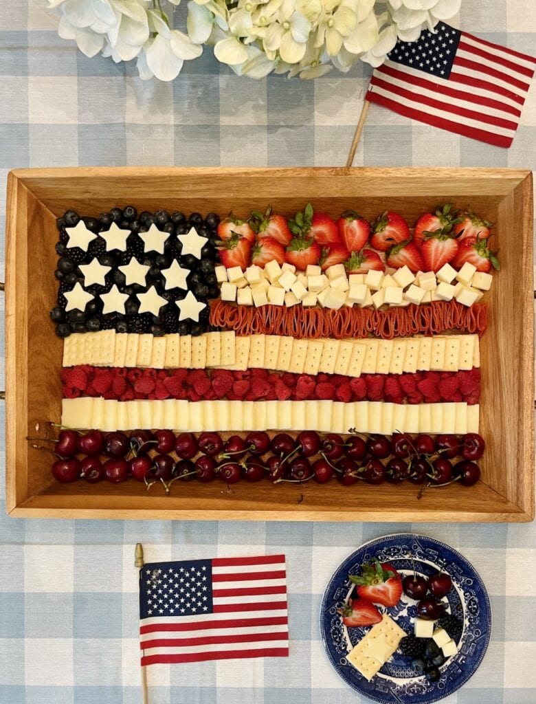 Flag Charcuterie Board on a wooden tray with red, white, and blue stripes of cherries, cheese, crackers, and berries arranged to look like the American flag, styled with white hydrangeas and American flags for a 4th of July celebration.