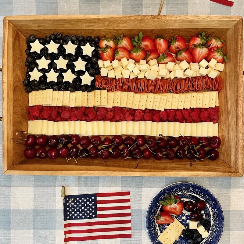 Flag Charcuterie Board on a wooden tray with red, white, and blue stripes of cherries, cheese, crackers, and berries arranged to look like the American flag, styled with white hydrangeas and American flags for a 4th of July celebration.