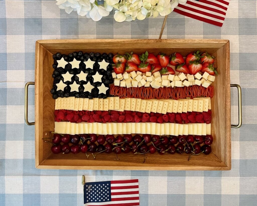Flag Charcuterie Board on a wooden tray with red, white, and blue stripes of cherries, cheese, crackers, and berries arranged to look like the American flag, styled with white hydrangeas and American flags for a 4th of July celebration.