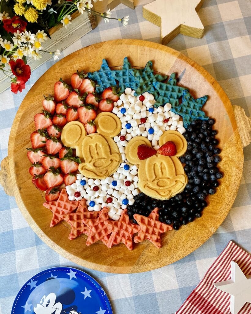 Mickey Patriotic Waffle Board with two golden Mickey waffles in the center surrounded by red and blue star waffles, fresh strawberries, blueberries, mini marshmallows, and M&Ms on a round wood tray for a Disney 4th of July brunch.