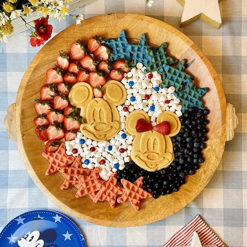Mickey Patriotic Waffle Board with two golden Mickey waffles in the center surrounded by red and blue star waffles, fresh strawberries, blueberries, mini marshmallows, and M&Ms on a round wood tray for a Disney 4th of July brunch.
