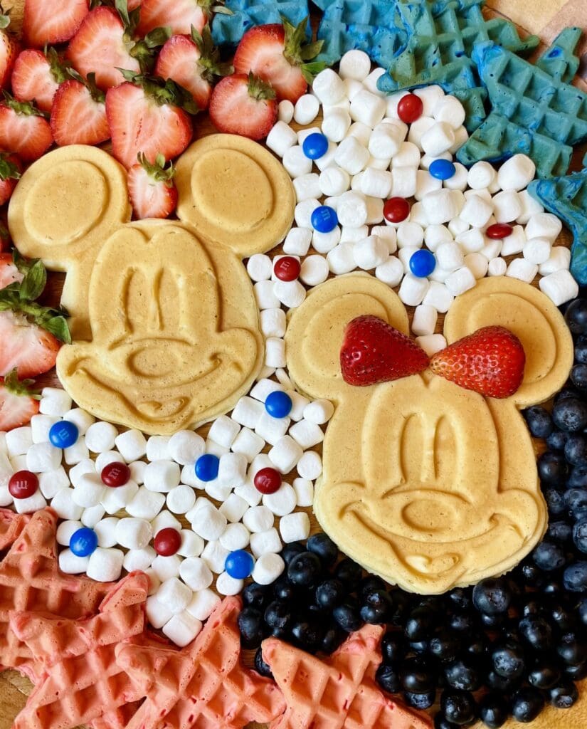 Mickey Patriotic Waffle Board with two golden Mickey waffles in the center surrounded by red and blue star waffles, fresh strawberries, blueberries, mini marshmallows, and M&Ms on a round wood tray for a Disney 4th of July brunch.