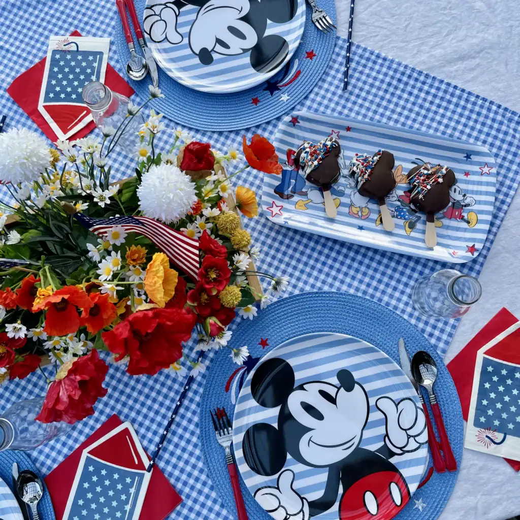 Star-print red napkin and rocket paper napkin layered at a patriotic Mickey place setting; navy star paper straws on a Disney patriotic gingham tablescape