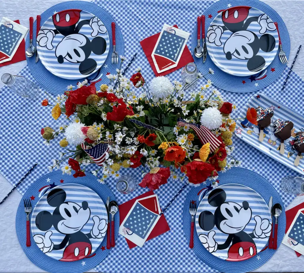 Retro Mickey striped dinner plate with red flatware and rocket napkin on a gingham patriotic table setting; wooden crate centerpiece with red poppies, white daisies, and American flags for a Disney 4th of July table