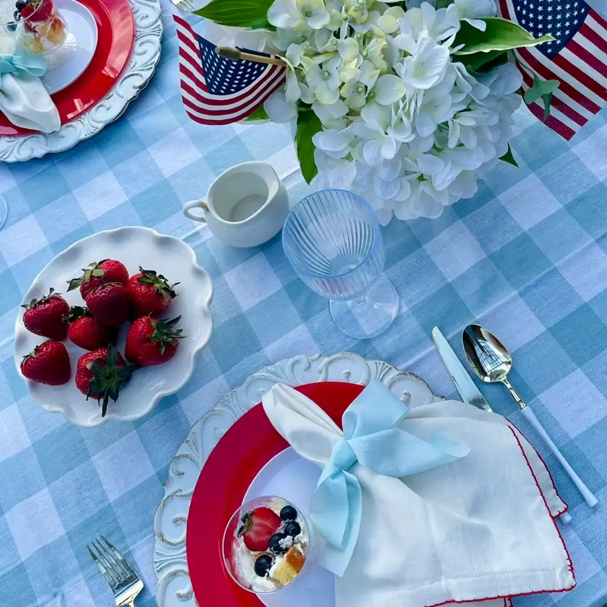 White cotton napkin with red blanket-stitch trim tied with light blue satin bow on a red and white patriotic place setting; white scrollwork charger with red disposable dinner plate and mini berry trifle cup — red white and blue cottage tablescape